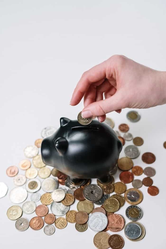 our-story Hand inserting coin into black piggy bank among scattered coins, symbolizing savings and financial planning.