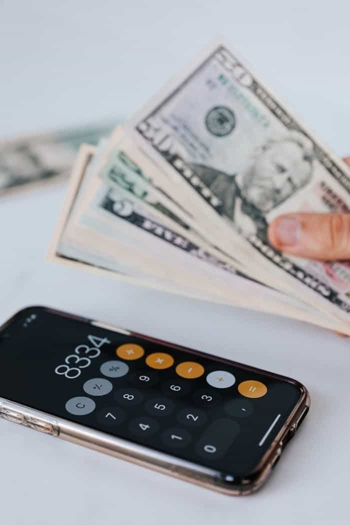 From above of crop anonymous person holding heap of American dollar near cellphone with calculator application representing numbers