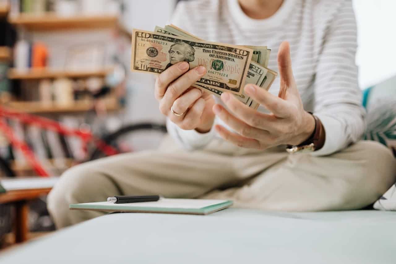 Man wearing casual clothes counts dollar bills while sitting on a sofa indoors.